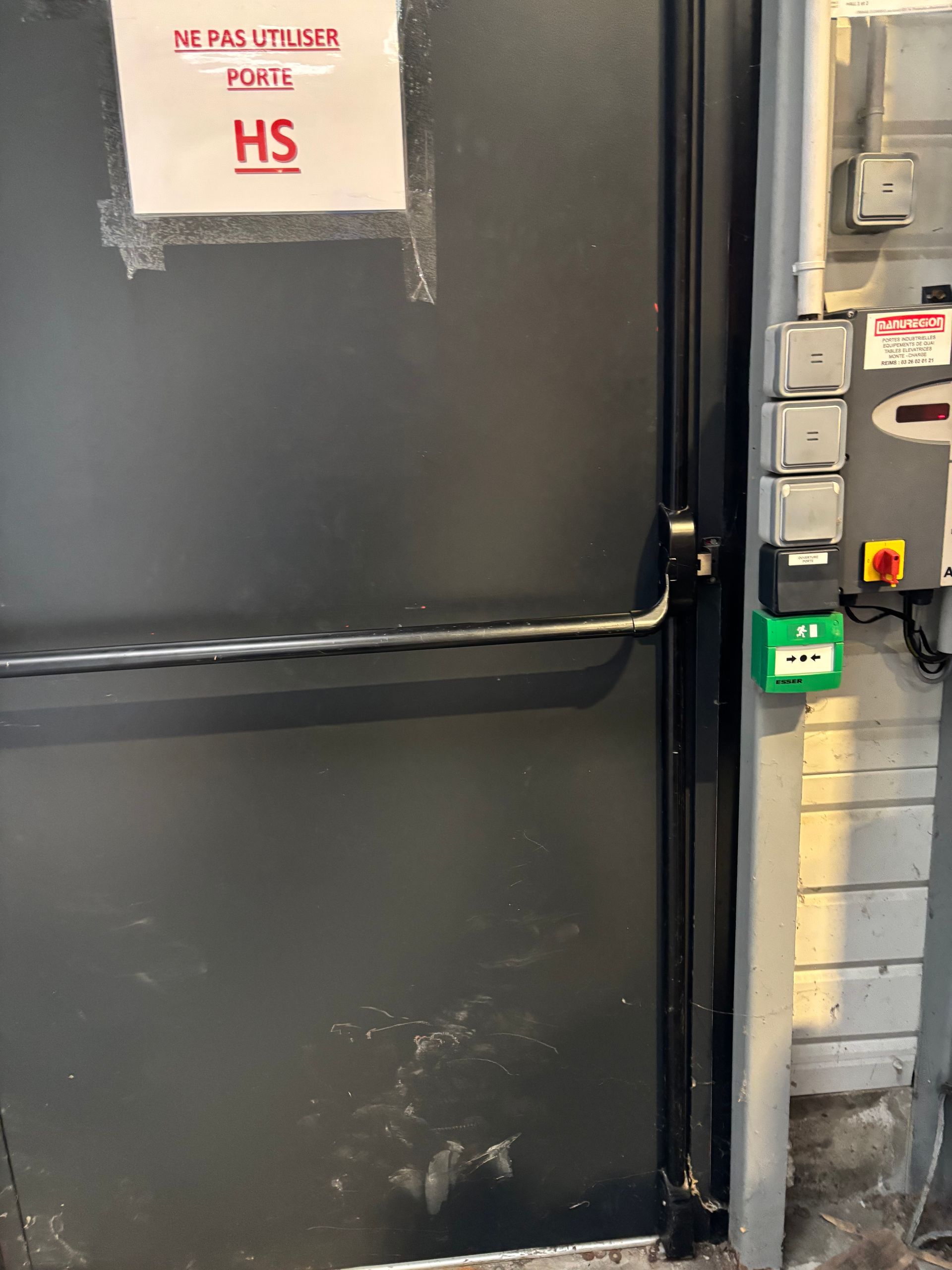Porte blindée industrielle noire à côté des panneaux de commande électriques dans une zone technique.
