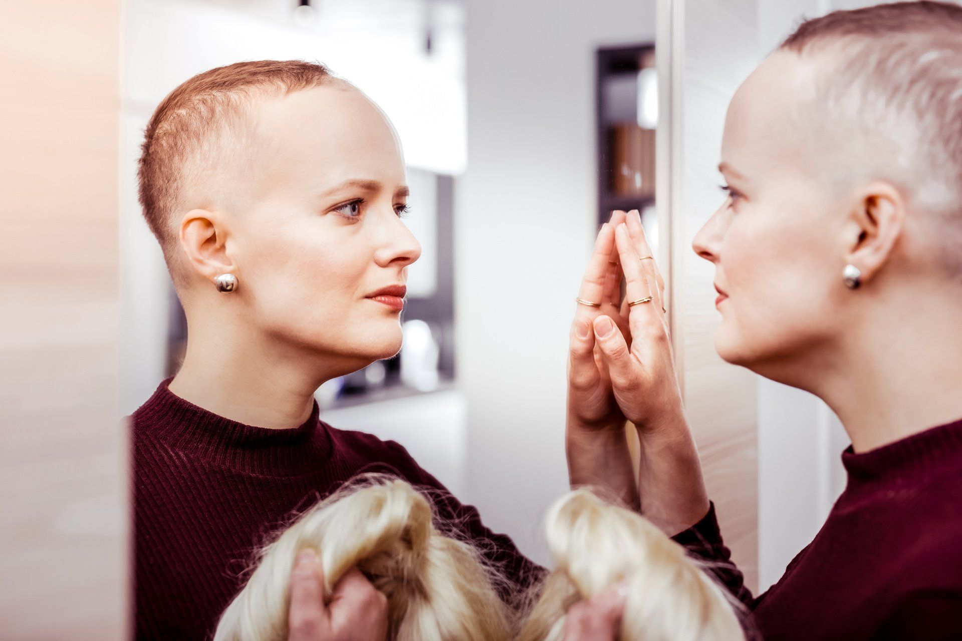 Femme se regardant dans le miroir, elle a perdu des cheveux et porte une perruque blonde dans la main