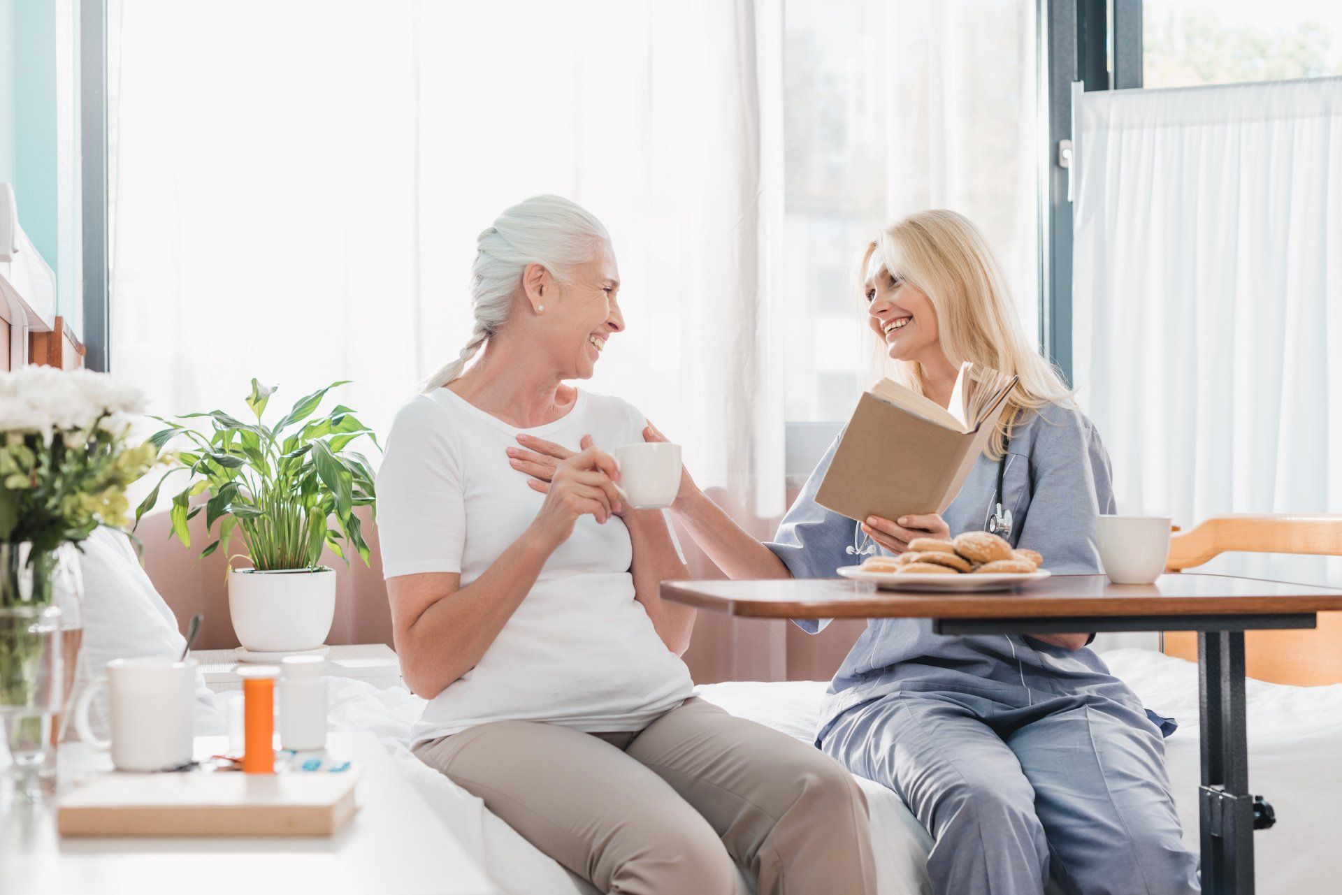 Femme âgée sur son lit médicalisé, avec soignante devant table de lit