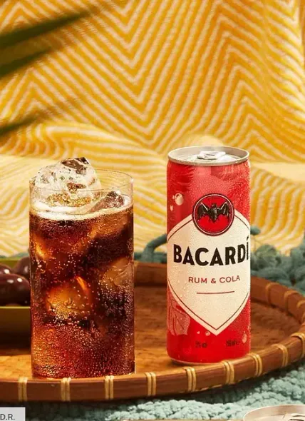 Una lata de ron bacardi con cola junto a un vaso de ron con cola.