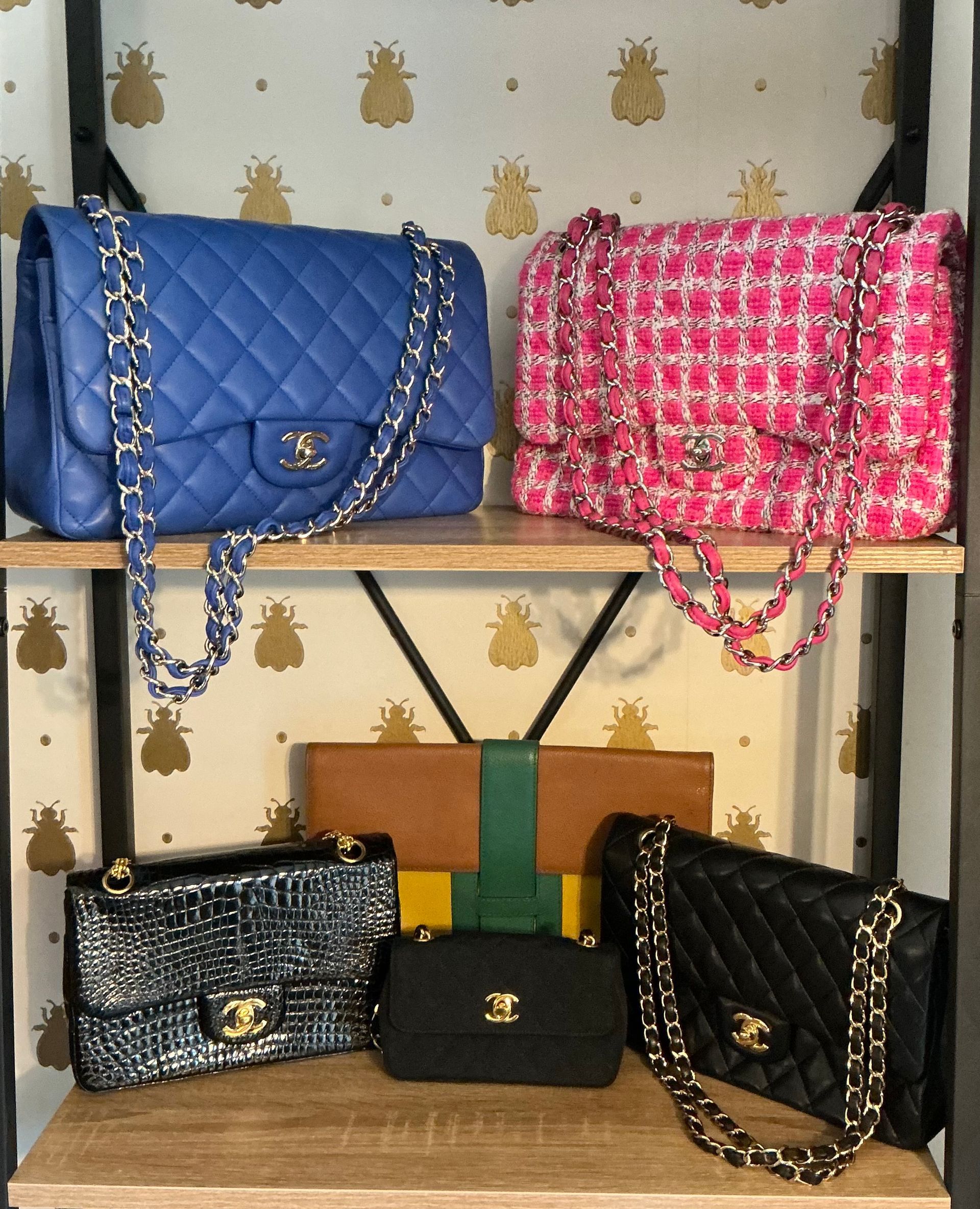 Sacs à main Chanel de couleurs et de styles variés, présentés sur des étagères en bois devant un fond à motifs.