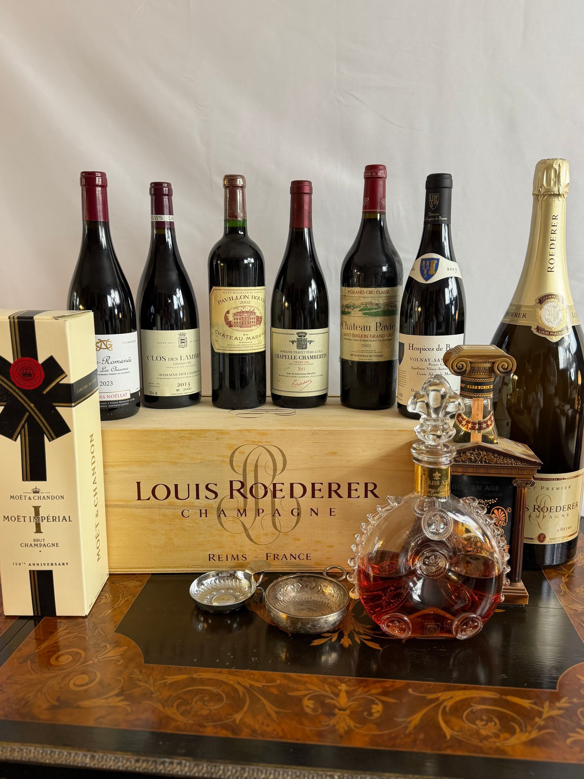 Plusieurs bouteilles de vin et une bouteille de champagne sont disposées sur une caisse en bois. Une boîte et une bouteille d'alcool sont également présentes.