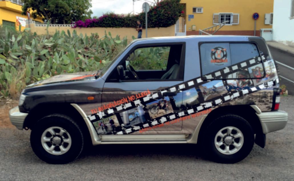 SUV azul y negra con diseño de tira de película estacionada en una calle cerca de plantas y un edificio amarillo.