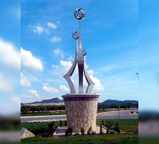 Una alta escultura moderna de metal con tres elementos esféricos se alza sobre un pedestal de piedra en una zona ajardinada al aire libre.