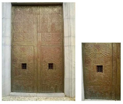 Un par de grandes puertas de bronce envejecido con intrincadas tallas en relieve y una pequeña ventana con barrotes en cada panel.
