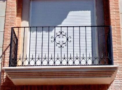 Una barandilla de balcón de metal negro con un diseño decorativo en forma de voluta en el centro, montada sobre una repisa de piedra contra una pared de ladrillo.