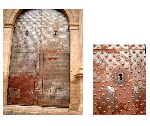 Una gran puerta metálica arqueada con una superficie oxidada y tachonada, mostrada en su totalidad, y un detalle en primer plano de su cerradura.