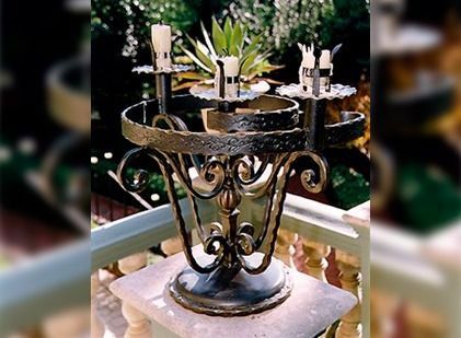 Candelabro ornamentado de metal oscuro con detalles de hierro forjado en espiral, que sostiene tres velas cilíndricas sobre un pedestal de piedra en el exterior.