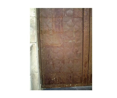 Una puerta de metal marrón, desgastada por el tiempo, presenta un patrón repetitivo en relieve que imita escamas de pez o vieiras, sobre una pared de piedra.