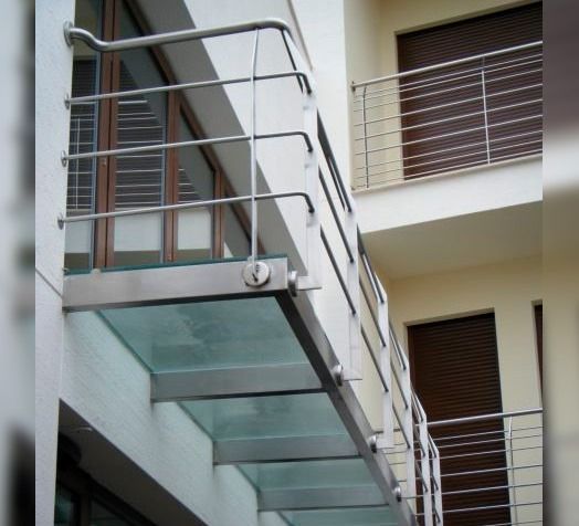 Un balcón moderno con suelo de cristal y barandilla de acero inoxidable, adosado a un edificio de varias plantas.