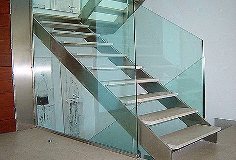 Escalera moderna con peldaños de piedra clara, largueros metálicos y barandilla de cristal transparente en un interior minimalista.