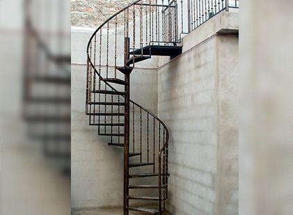 En un espacio interior, una escalera de caracol metálica con barandillas oscuras se alza contra una pared lisa de bloques de color claro.