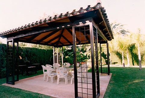 Un cenador negro con estructura metálica y techo de tejas se encuentra en un patio pavimentado en un jardín trasero con césped y muebles de comedor blancos.