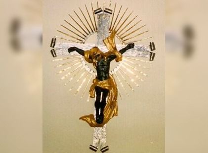 Un crucifijo de cuerpo negro con drapeado dorado, colocado sobre una cruz plateada adornada con un diseño de rayos de sol radiantes.