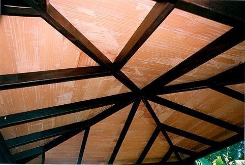 Vista desde un ángulo bajo del techo de un cenador de madera, con vigas oscuras que irradian desde el centro sobre paneles de madera de color claro.