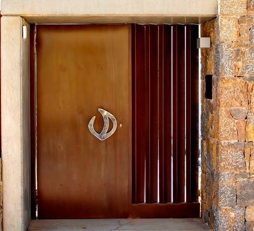 Una moderna puerta metálica con un tirador plateado de diseño escultural, integrada en una pared de piedra junto a una mampara oscura de listones verticales que proporciona privacidad.