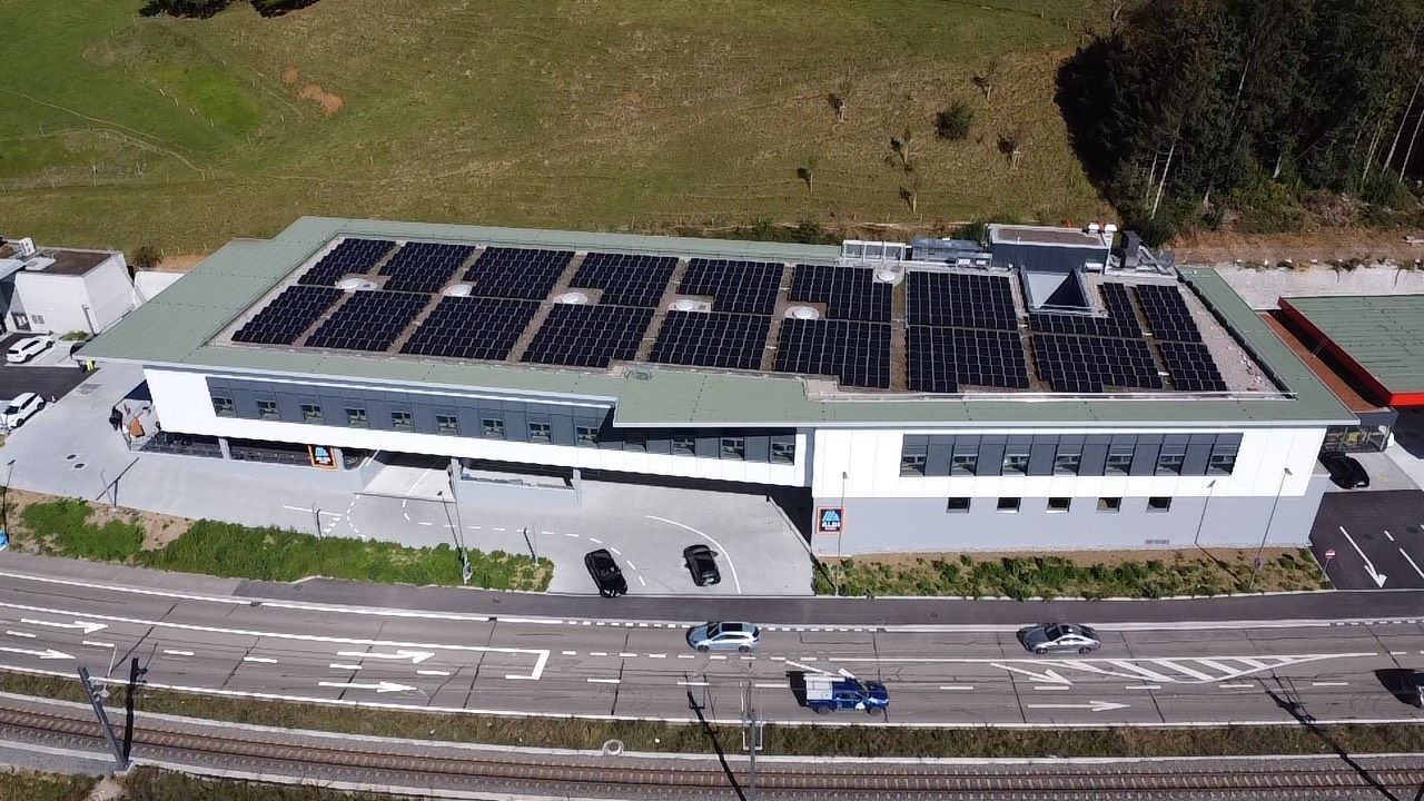 Gebäude mit Solarmodulen auf dem Dach, neben einer Straße und Bahngleisen.