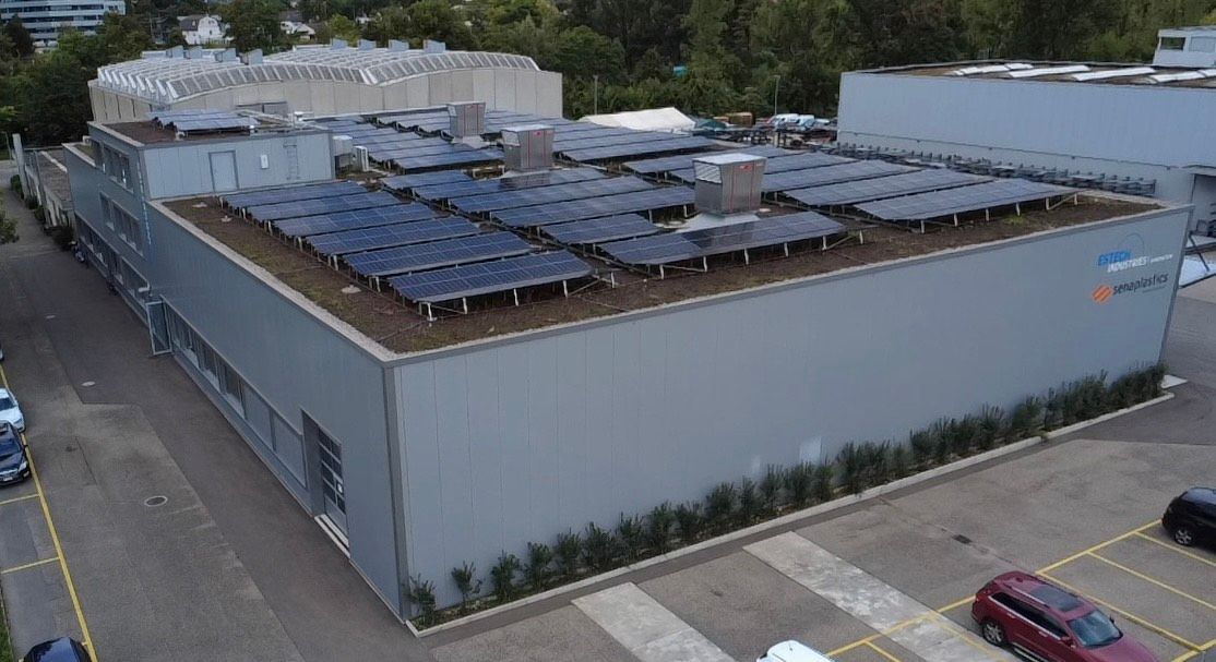 Gebäude mit Solarmodulen auf dem Dach, umgeben von geparkten Autos und Bäumen.