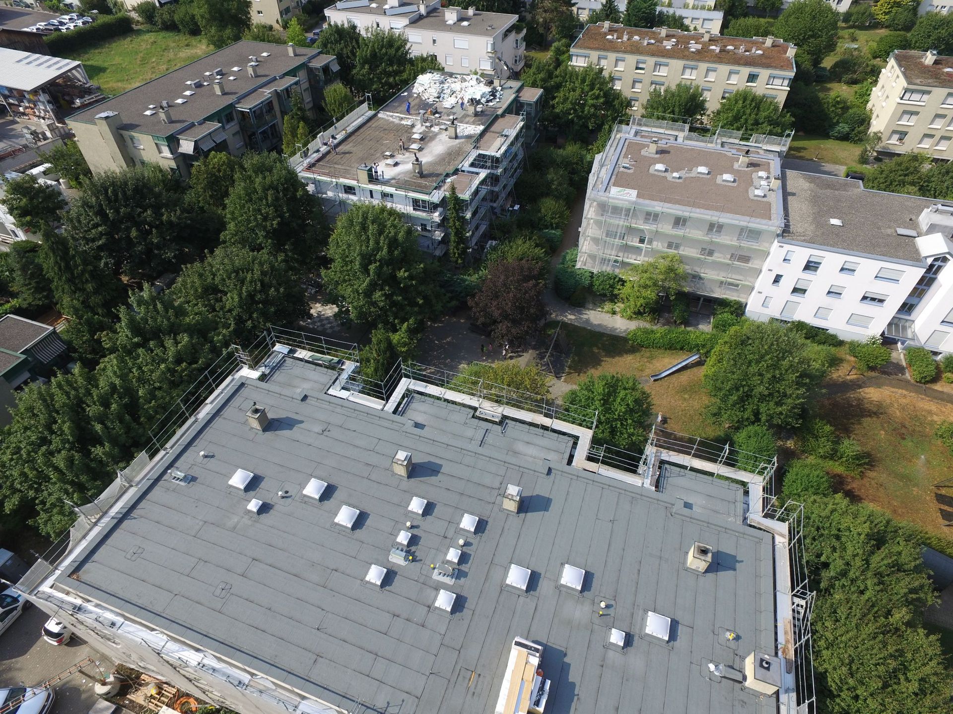 Luftaufnahme von Wohnhäusern mit dunklen Dächern, Bäumen und grünen Grasflächen. Sonniger Tag.