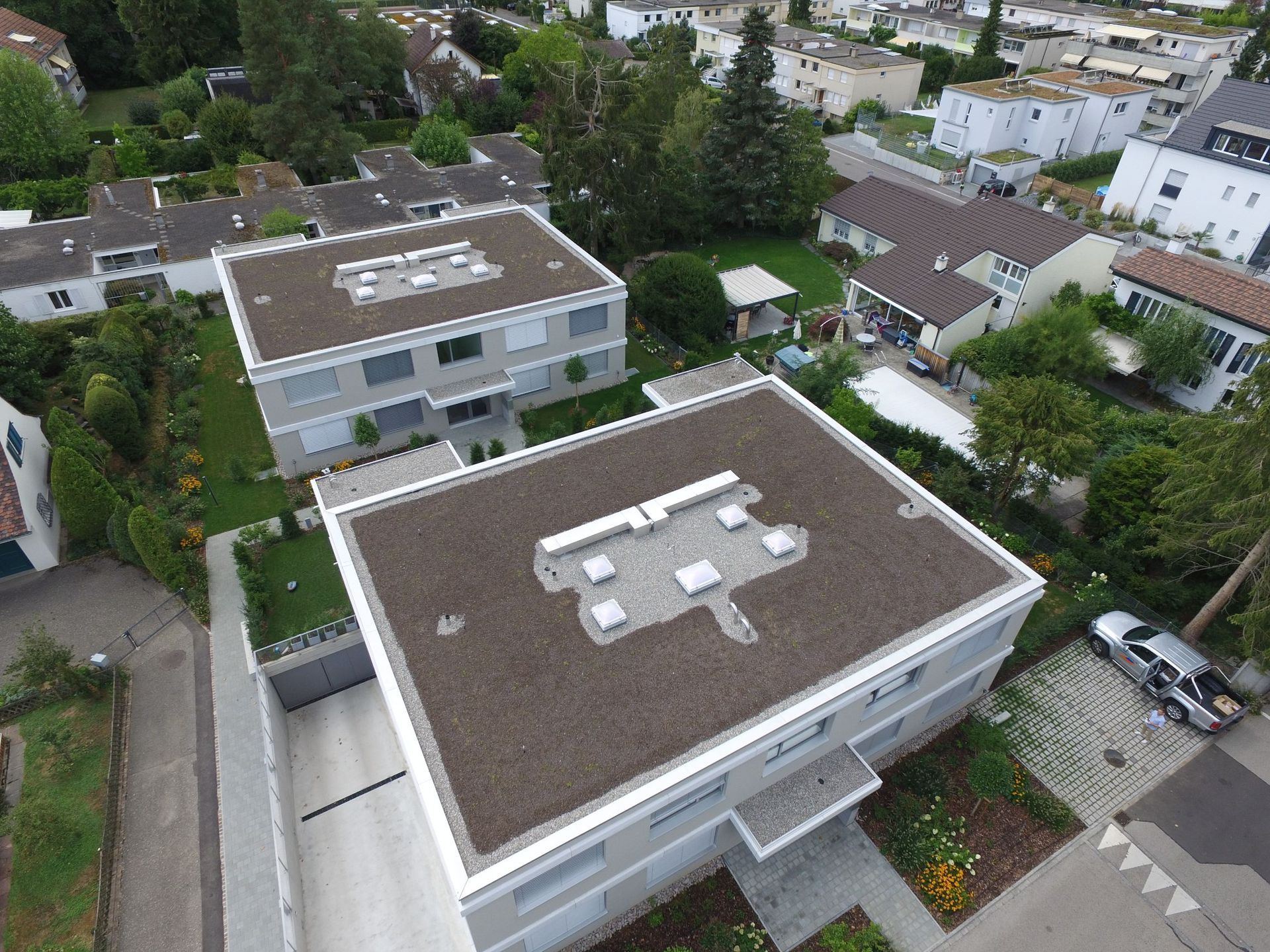 Luftaufnahme von zwei modernen Gebäuden mit flachen braunen Dächern, umgeben von Grün und anderen Häusern.