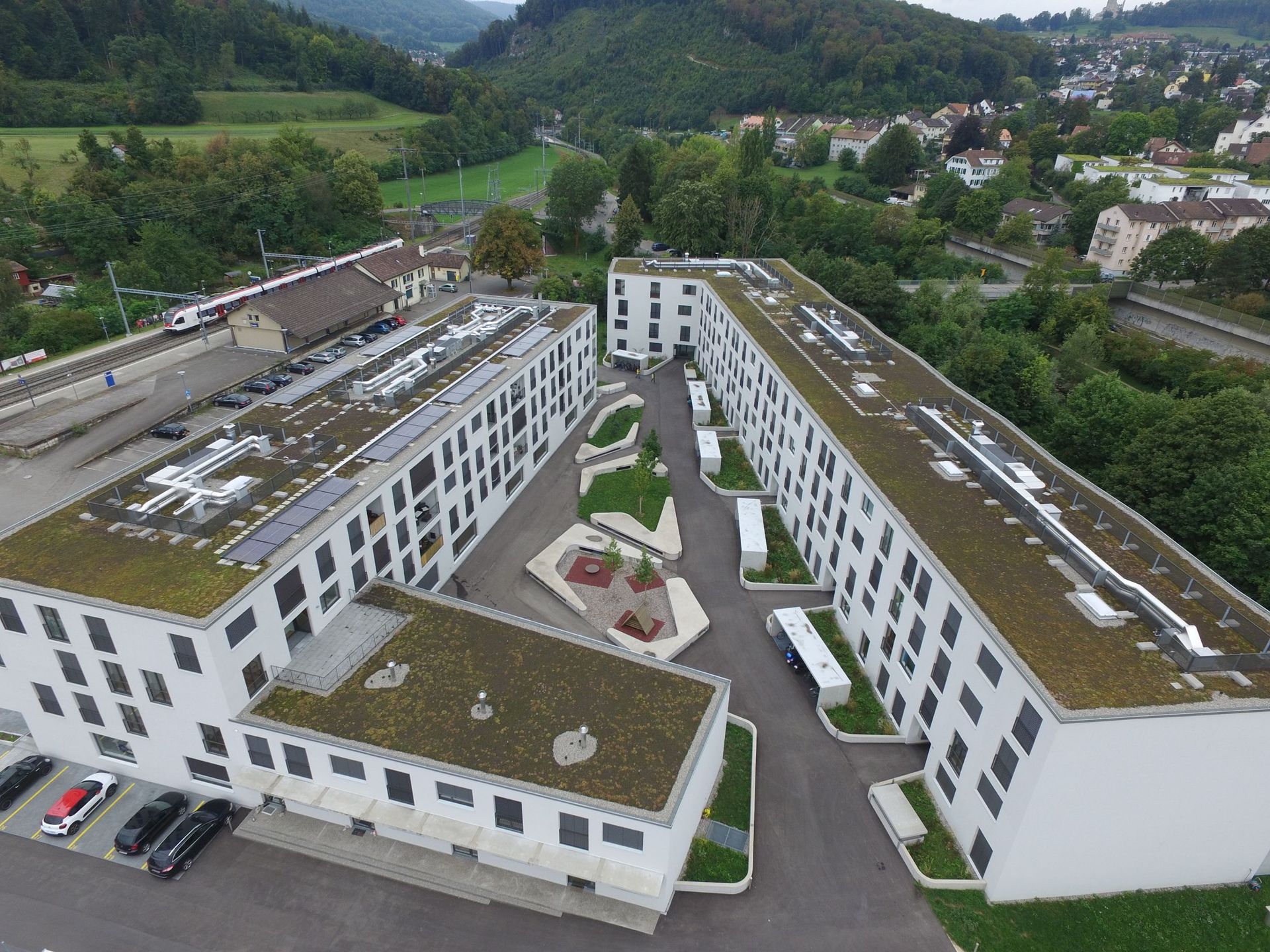 Luftaufnahme moderner weißer Wohnhäuser mit grünen Dächern, eingebettet in der Nähe eines Bahnhofs und eines Hügels.