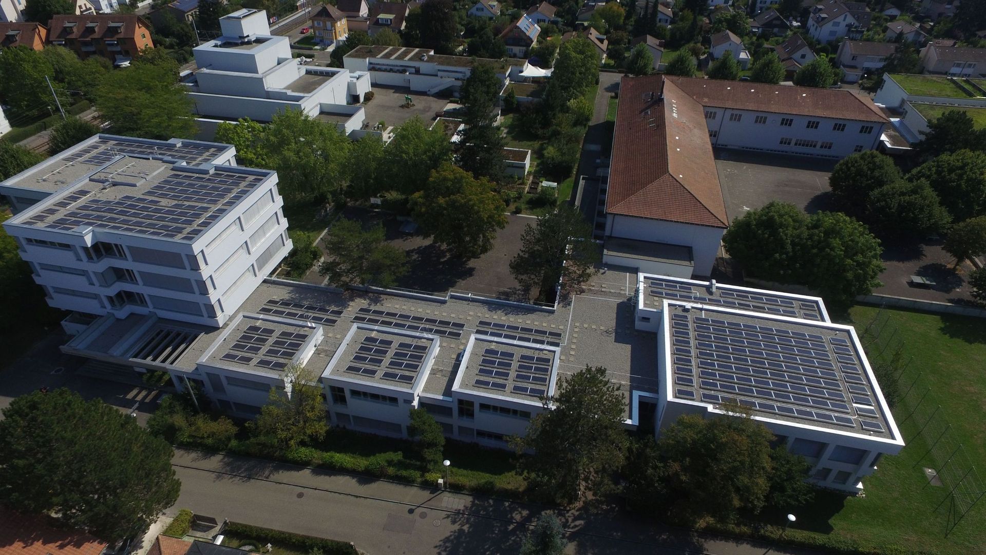 Luftaufnahme von Gebäuden mit Solarmodulen auf den Dächern, umgeben von Bäumen und grünen Rasenflächen.