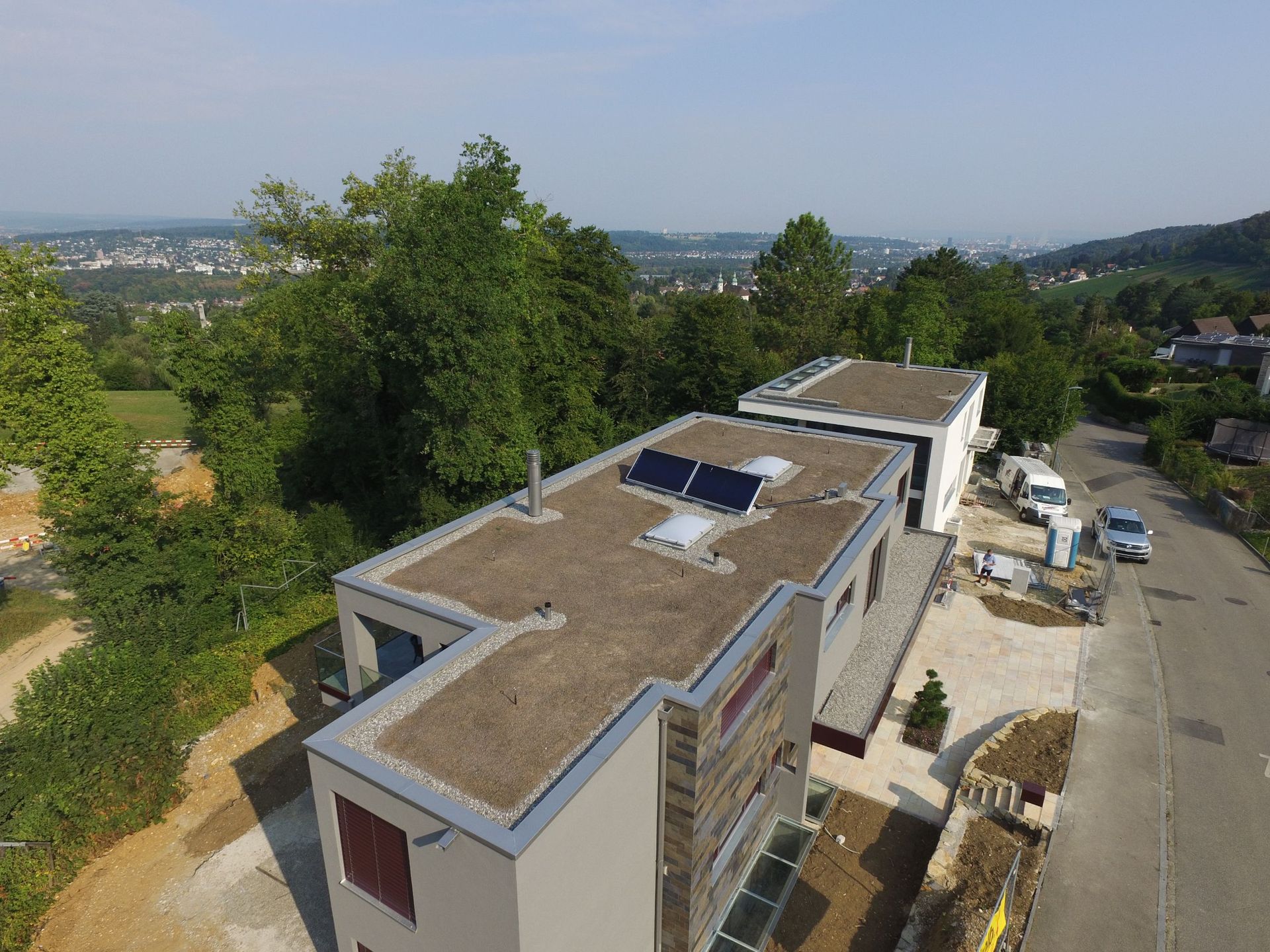 Modernes Haus mit Solarmodulen auf einem Flachdach, Bäumen und einer entfernten Stadtlandschaft.
