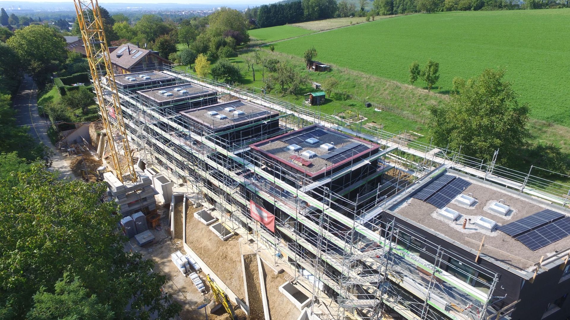 Baustelle mit Gerüst, Kran und langem Gebäude mit Flachdach und Solarmodulen.