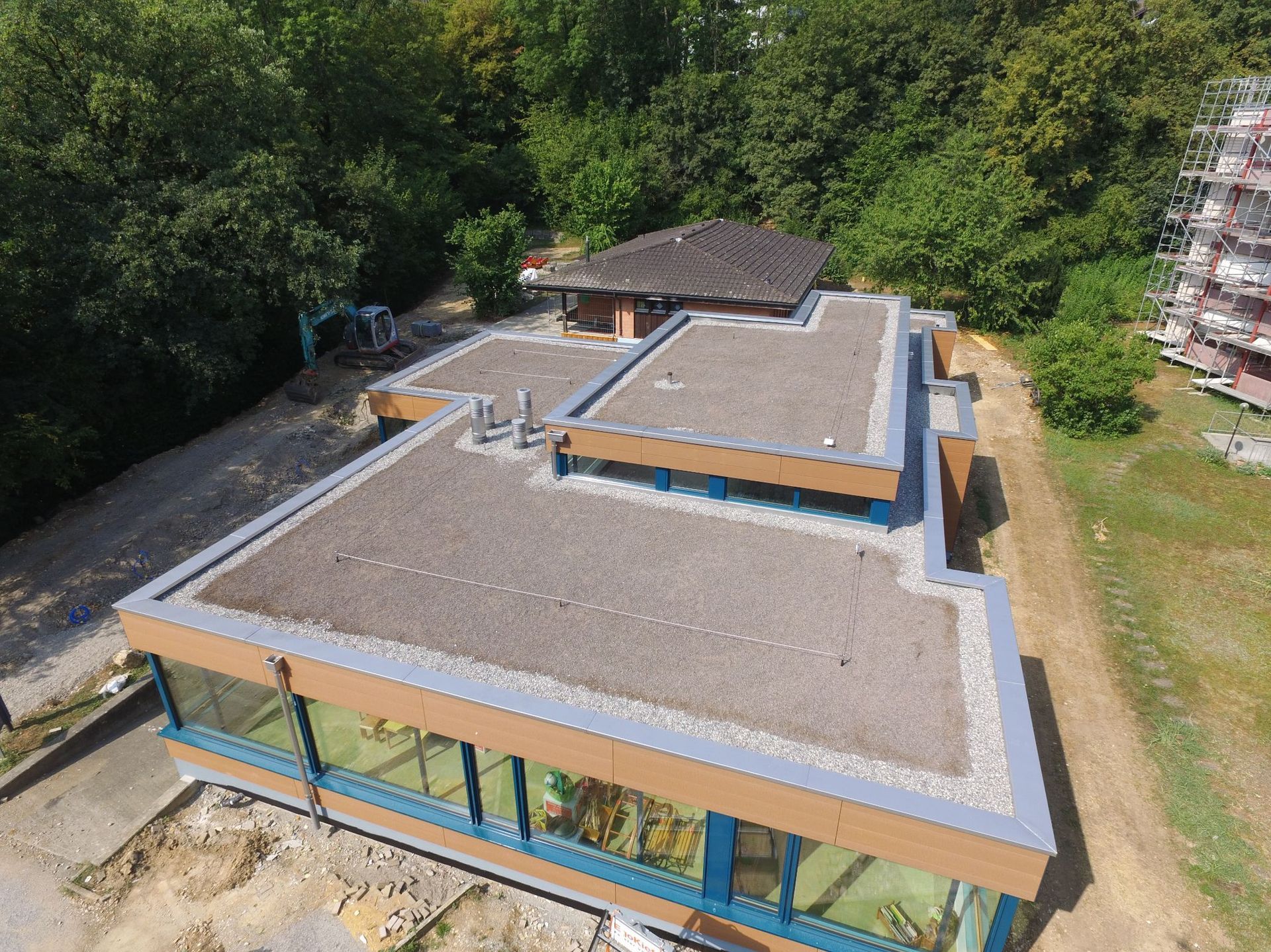 Luftaufnahme eines modernen Gebäudes mit Flachdach und Glasfenstern, umgeben von Bäumen, Baustellen und Erde.