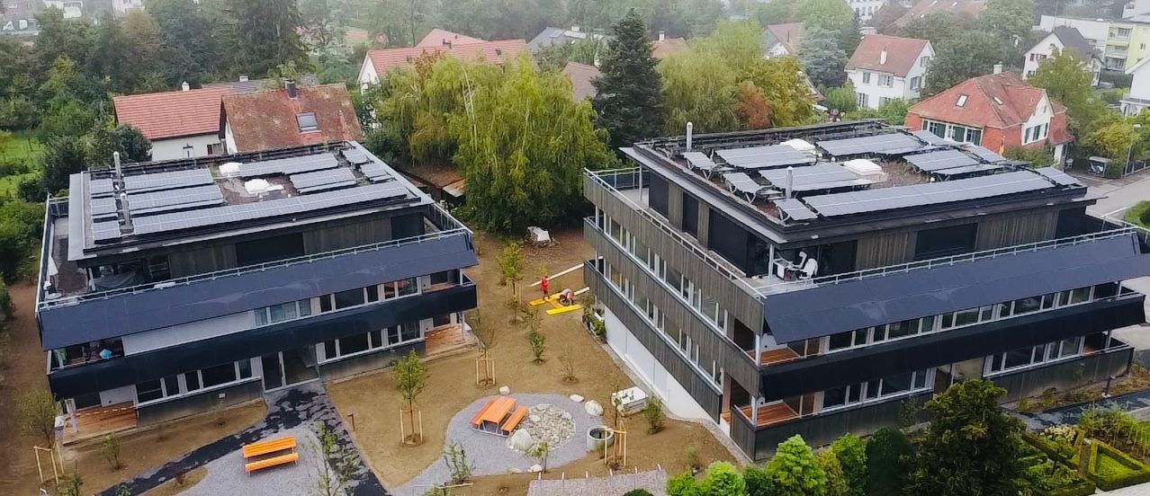 Zwei moderne Mehrfamilienhäuser mit Solarmodulen auf den Dächern in einem Wohngebiet.
