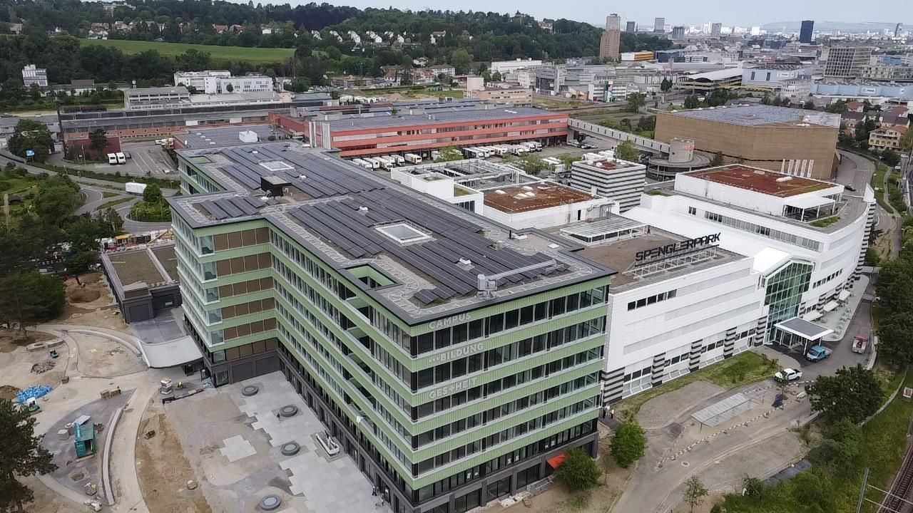 Luftaufnahme moderner Gebäude mit Solarmodulen, umgeben von Stadt und Grün.