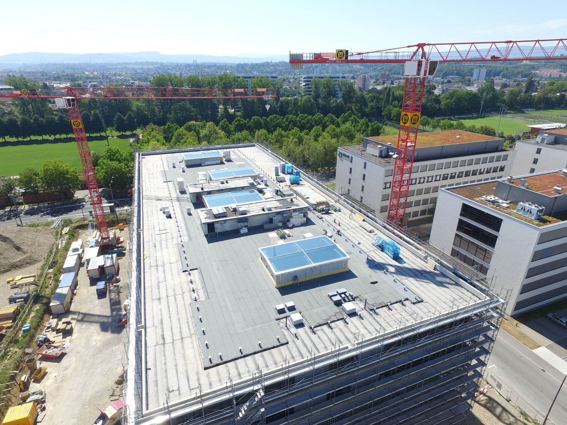 Baustelle mit dem Flachdach eines hohen Gebäudes, roten Kränen, weißen Gebäuden und einer grünen Wiese.