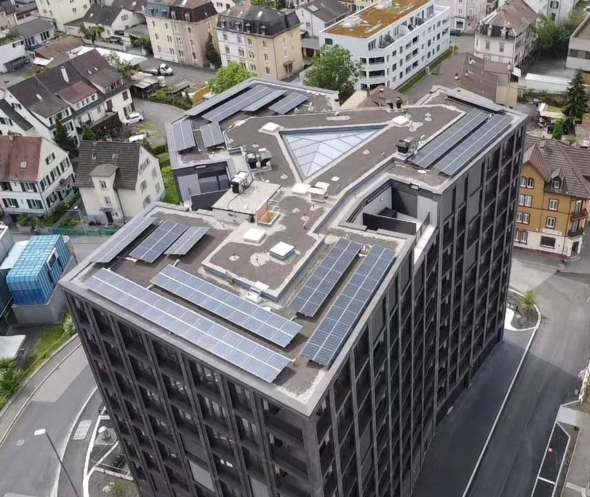 Hochhaus mit Solarmodulen auf dem Dach. Umgeben von Bäumen und anderen Gebäuden in einer Stadt.