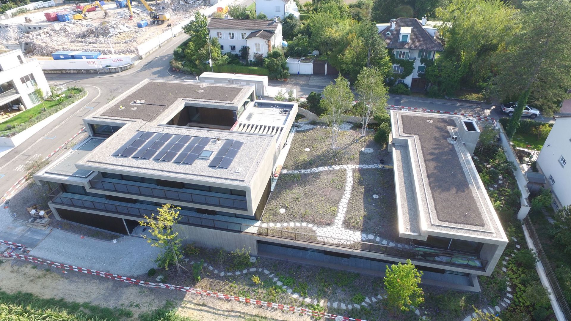 Luftaufnahme moderner Gebäude mit Sonnenkollektoren und Gründächern. Baustelle und Bäume im Hintergrund.