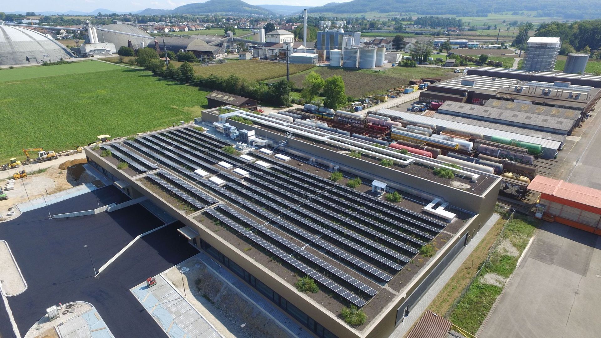 Solarmodule auf einem Gebäudedach, im Hintergrund Industriegebiet und Felder.