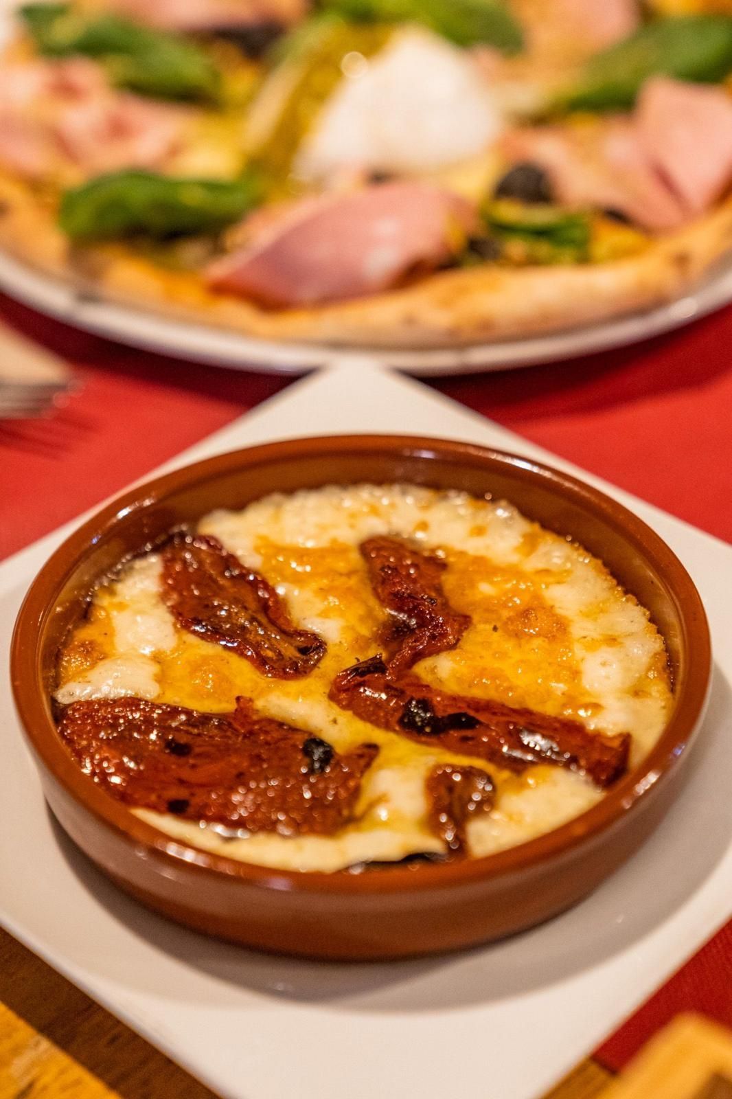 Plato de queso al horno con tomates secados al sol, en un recipiente de cerámica marrón sobre un plato blanco; pizza en el fondo.