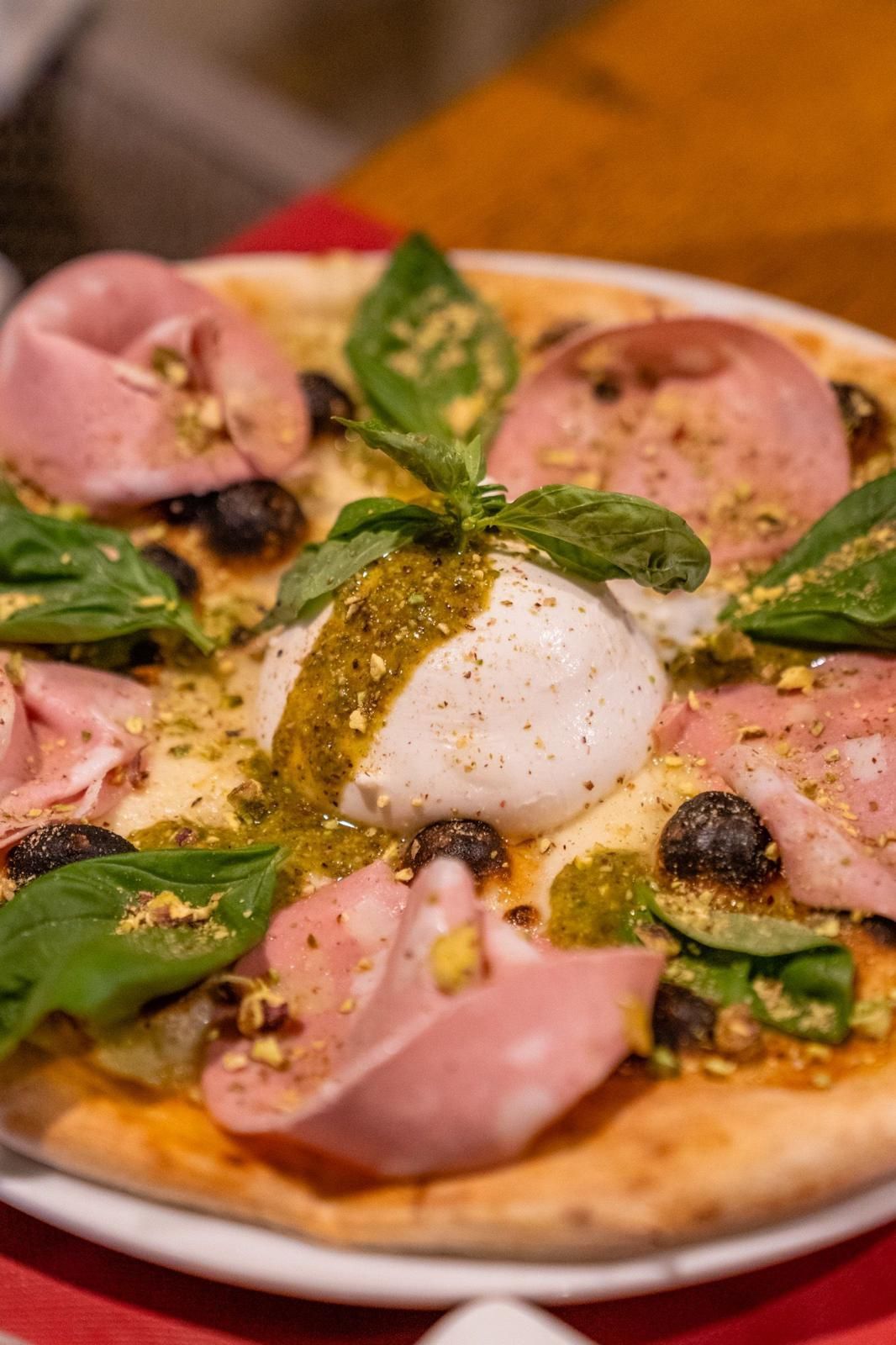 Pizza cubierta con queso burrata, jamón, aceitunas, albahaca y migas de pistacho.
