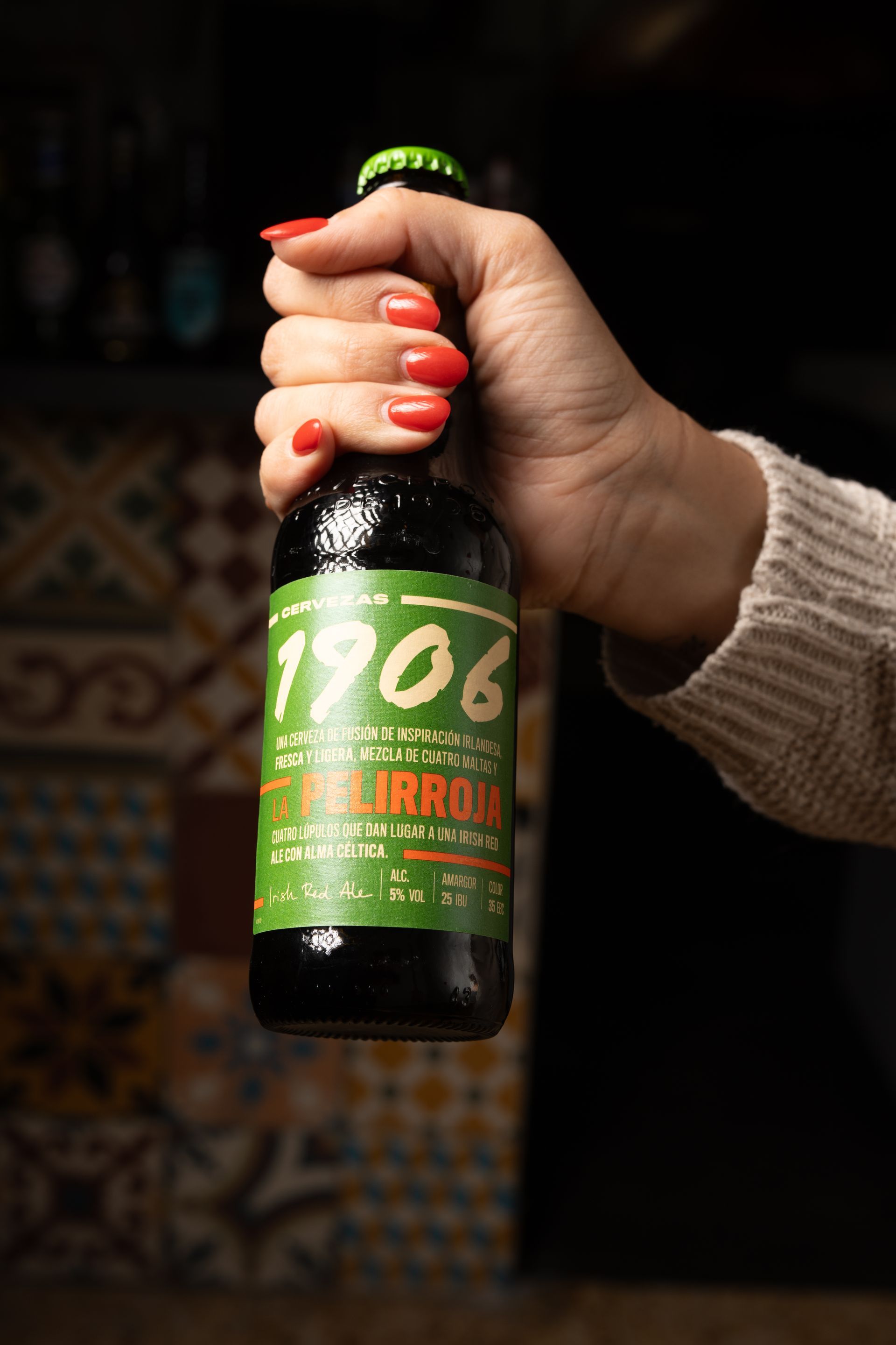 Mano sosteniendo una botella de cerveza de 1906 con etiqueta verde y blanca contra un fondo de bar de azulejos.