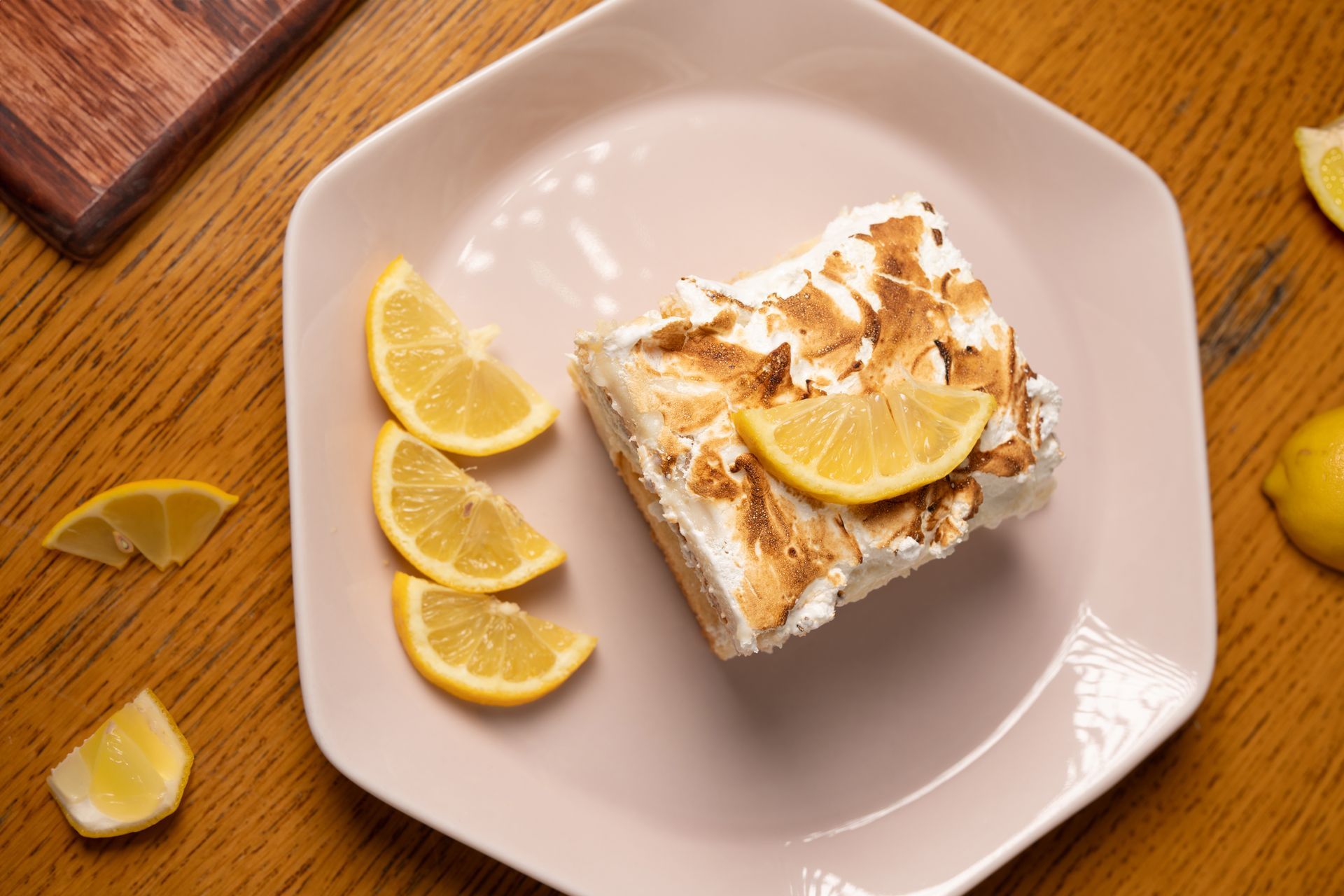 Rebanada de postre de merengue de limón con gajos de limón en plato rosa, mesa de madera.