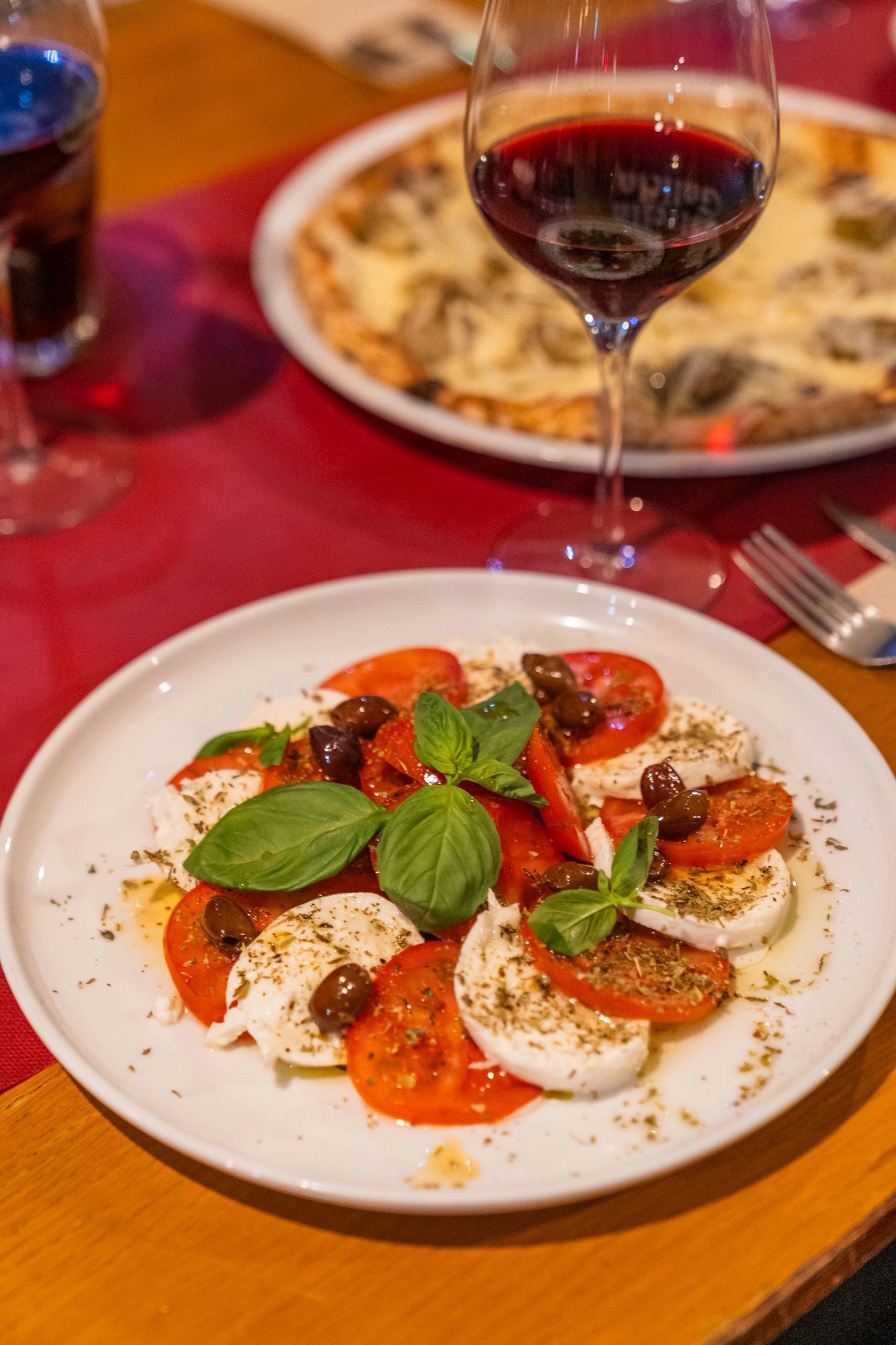 Ensalada caprese con vino tinto, pizza de fondo, sobre mantel rojo.