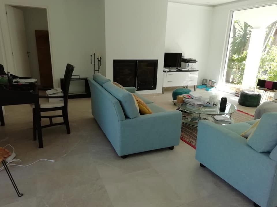 Sala de estar luminosa con sofás azul claro, televisor, mesa de comedor y ventanas que dejan entrar la luz del sol.