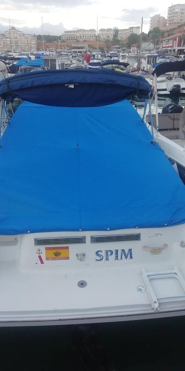 Popa de barco blanca con cubierta azul brillante y bandera española en un puerto, edificios de la ciudad al fondo.