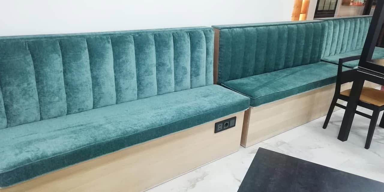 Banco tapizado en color verde azulado con base de madera en una moderna sala de espera interior, junto a una silla negra.