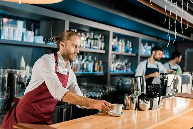 Camarero con delantal marrón colocando una taza en la barra; otros dos trabajan detrás de él.