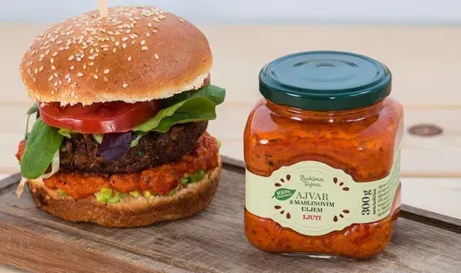 Hamburguesa con salsa roja y un tarro de salsa ajvar sobre una tabla de madera.