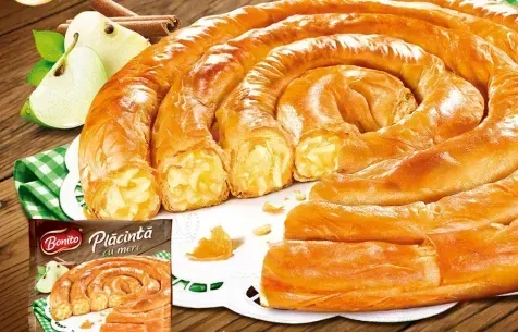 Pastel en forma de espiral con corteza dorada, cortado para revelar un relleno, junto a manzanas y ramas de canela.