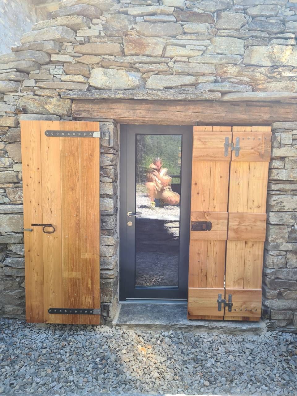 Edificio in pietra con una porta incorniciata da persiane in legno. La porta ha un pannello di vetro che riflette una figura.