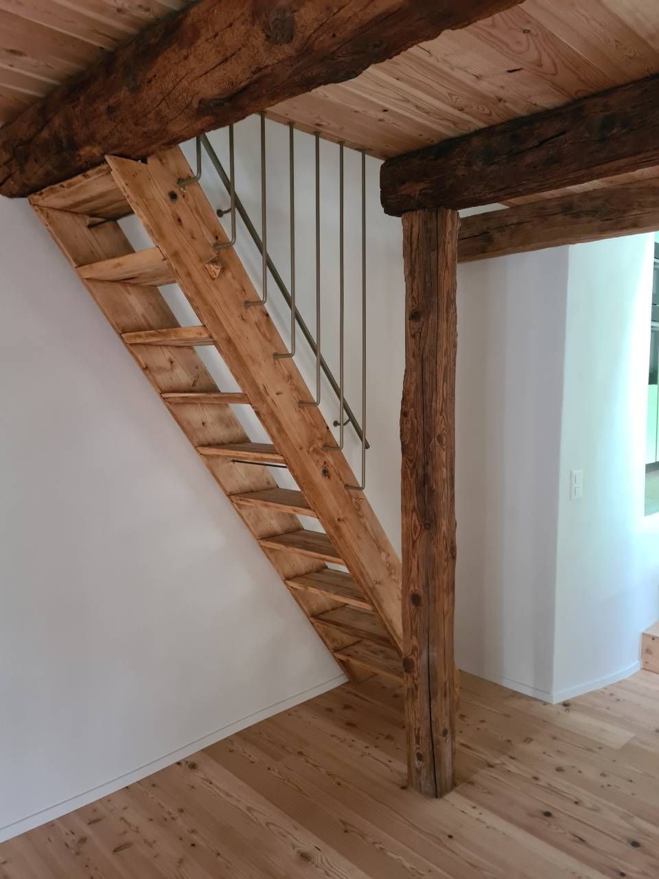 Scala in legno con ringhiera in metallo e travi a vista in una stanza con pavimenti in legno chiaro e pareti bianche.