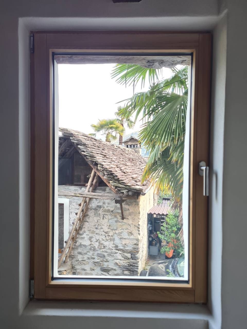 Vista dalla finestra di un edificio in pietra con tetto di tegole, palme e giardino.