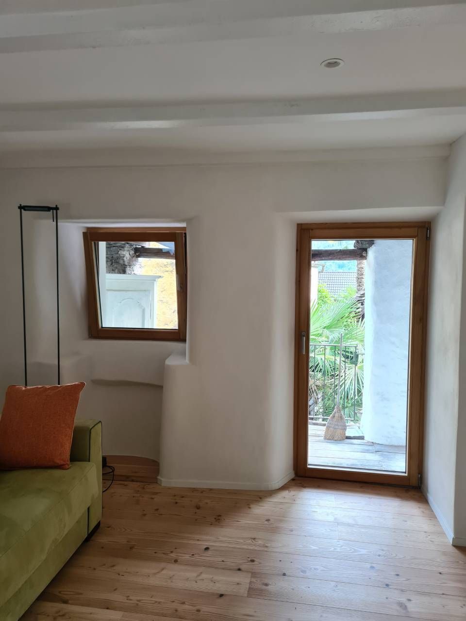 Stanza interna con divano verde, pavimento in legno, finestra e porta che conduce a un'area esterna.